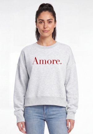 Frau mit dunklen, zurückgebundenen Haaren, die einen hellgrauen Pullover mit rotem Text "Amore." und blaue Jeans trägt, stehend vor weißem Hintergrund.