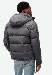 Chaqueta de plumas gris con capucha, con diseño acolchado, bordado del logo en el hombro y puños elásticos. Combinada con jeans decolorados.