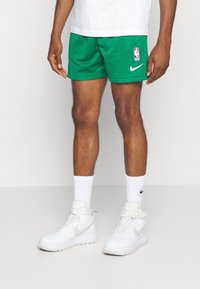 Uomo che indossa pantaloni da basket NBA verdi, calze bianche Nike e scarpe alte bianche Nike, in piedi contro uno sfondo semplice.