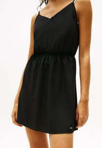 Schwarzes, ärmelloses Kleid mit taillierter Waist, einem leichten V-Ausschnitt, Spitzenbesatz und einem kleinen Logopunkt nahe dem Saum. Leichter Stoff.