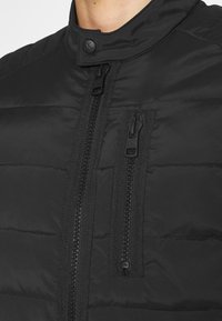 Black padded jacket with vertical front and chest zippers, a snap button collar, and "ck" logo on zipper pulls and button.