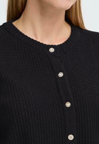 Svart strikket cardigan med ribbet hals, seks dekorative metallknapper og et teksturert, koselig design.