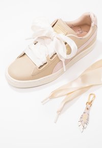 Baskets en cuir beige avec des accents roses, grandes lacets en satin blanc, accessoires en or, et un porte-clés décoratif en forme de feuille. Semelle texturée.