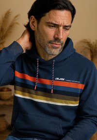 Marineblauwe hoodie met meerkleurige horizontale strepen in rood, blauw, groen en wit. Voorzien van een capuchon met trekkoord en een vo zak.