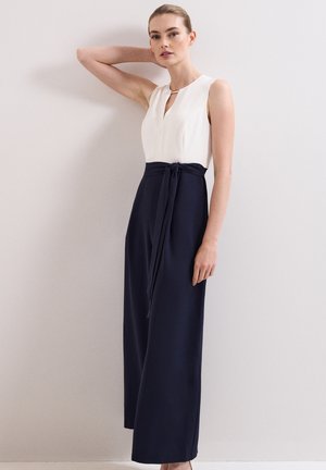 Vrouw draagt een mouwloze witte top met keyhole-halslijn en lange marineblauwe broek met gestrikte taille, staand tegen een effen lichte muur.