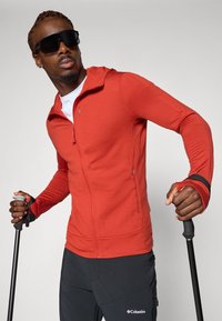 Rode zip-up jas met lange mouwen en duimgaten, gecombineerd met zwarte outdoor broek. Zwarte wandelstok vastgehouden in een gewatteerde hand.