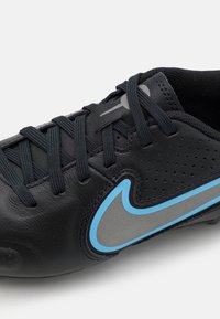 Botín de fútbol de cuero negro con acentos grises y un logo de swoosh azul. Presenta una superficie texturizada, perforaciones y cordones negros.