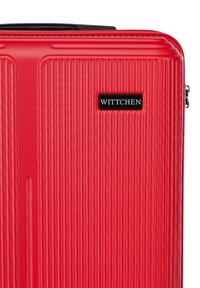 Valise rigide rouge avec texture à rainures verticales, dotée d'une plaque de logo noire et d'une fermeture éclair. Matériau durable, design compact.