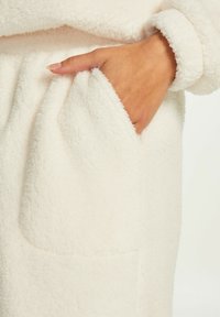 Bílá, huňatá tkanina s jemnou texturou; má viditelnou kapsu na boku a manžetu, která končí na zápěstí.