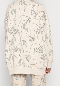 Krémová mikina s opakujícím se vzorem rukou v černých konturách a tyrkysovými akcenty na konečcích prstů. Uvolněný střih a jemná textura.