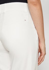 Białe spodnie o gładkiej fakturze, z tylną kieszenią i małym metalowym akcentem w postaci logo. Pas jest płaski i strukturalny.