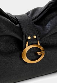 Sac à main en cuir noir à surface texturée, avec une boucle circulaire dorée et le logo « GUESS ». Design doux et plissé en haut.