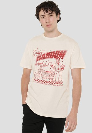 Young man with curly hair wearing a beige Duke Caboom t-shirt featuring a cartoon motorcycle and character illustration.
