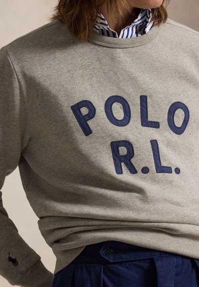 Personne portant un sweat gris avec le texte « POLO R.L. », une chemise rayée bleu marine, bleue et blanche à col, et un pantalon bleu marine.