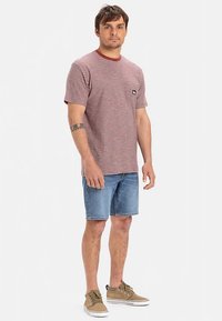 Červené a biele pruhované tričko s krátkym rukávom a okrúhlym výstrihom, skombinované s modrými džínsovými kraťasmi a svetlohnedými teniskami. Na tričku je viditeľné logo značky.