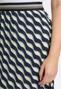 Jupe à motifs ondulés en marine, crème et vert olive. Présente une taille noire avec des accents métalliques. Tissu léger avec une texture lisse.