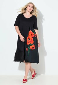 Frau im schwarzen Midikleid mit orangefarbener Mohnblumen-Print auf einer Seite und roten offenen Schuhen mit Absatz, stehend vor weißem Hintergrund.
