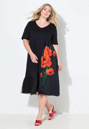 Kvinna i svart midiklänning med apelsinpoppy-mönster på ena sidan och röda öppna klackar, stående mot vit bakgrund.