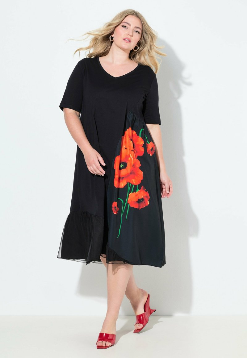 Frau im schwarzen Midikleid mit orangefarbener Mohnblumen-Print auf einer Seite und roten offenen Schuhen mit Absatz, stehend vor weißem Hintergrund.