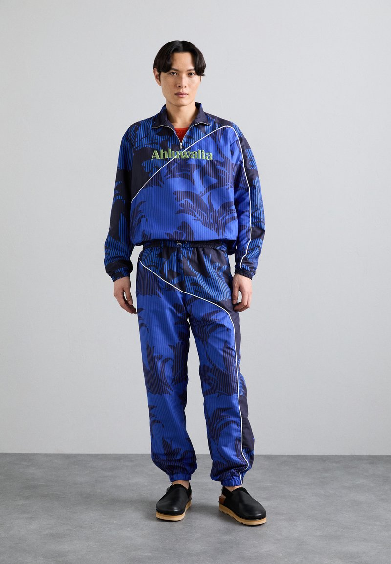 Trainingspak in het blauw met een grafisch bloemenpatroon, voorzien van elastische manchetten en een groen logo. Gepaard met zwarte schoenen met houten zolen.