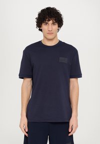 Sötétkék pamut t-shirt kerek nyakkivágással, rövid ujjakkal és téglalap alakú folttal a mellen. Szabott fazon.