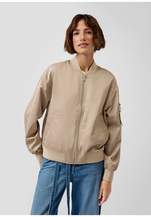 Frau mit kurzen braunen Haaren, die eine beige Bomberjacke mit Reißverschluss und blaue Jeans trägt und vor einem schlichten hellen Hintergrund steht.