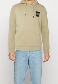 Sudadera beige con un bolsillo canguro y capucha ajustable con cordón. Cuenta con un pequeño parche de logo negro en el pecho y puños acanalados.