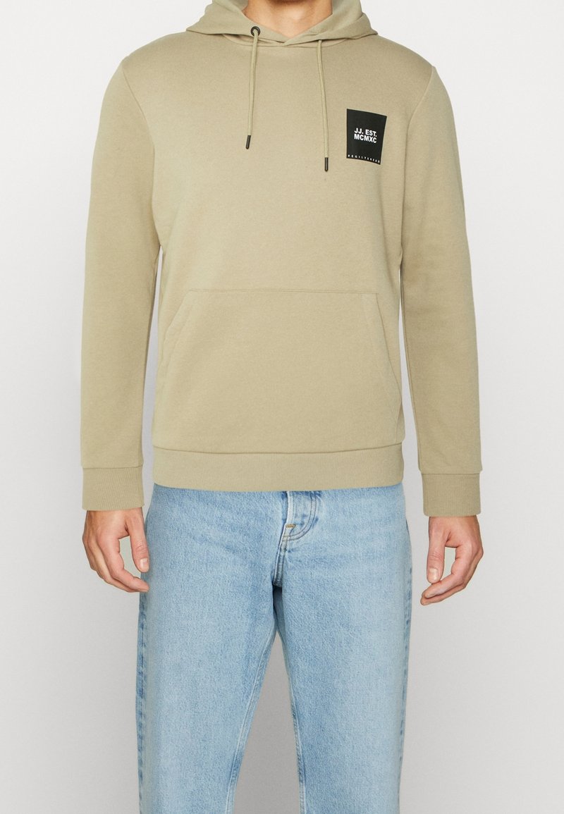 Sudadera beige con un bolsillo canguro y capucha ajustable con cordón. Cuenta con un pequeño parche de logo negro en el pecho y puños acanalados.