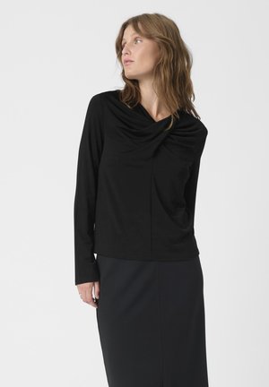 Black long-sleeve top with a draped cowl neckline, made of soft fabric. Paired with a black skirt, the outfit is clean and monochromatic.