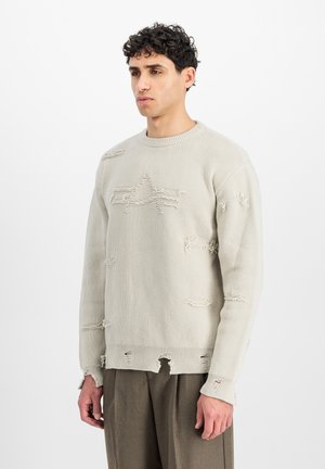 Ung mand med kort krøllet hår iført en beige slidt striktrøje og brune bukser, stående mod en ensfarvet hvid baggrund.
