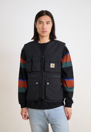 Czarna ocieplana kamizelka z wieloma przednimi kieszeniami; posiada zamek błyskawiczny, wykończenie dołu i logo Carhartt. Nosi się ją na czarnym swetrze w wielokolorowe paski.