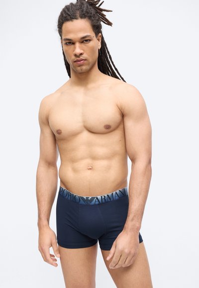 Εμφανίσιμος άνδρας με μακριά ράστα, φορώντας σκούρα μπλε μποξεράκια Emporio Armani, στέκεται μπροστά σε απλό φωτεινό φόντο.