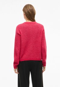 Rdeča pletena majica z okroglem vratnim izrezom, dolgimi rokavi in sproščenim krojem. Ribrana struktura in rob, v kombinaciji s črnimi hlačami.