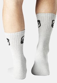 Jambes portant des chaussettes mi-mollet grises claires avec des motifs de visages souriants noirs sur les côtés externes, présentées de dos sur un fond blanc.