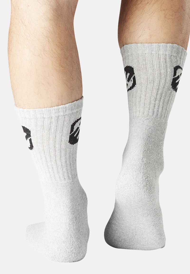 Jambes portant des chaussettes mi-mollet grises claires avec des motifs de visages souriants noirs sur les côtés externes, présentées de dos sur un fond blanc.
