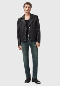 Černá kožená motocyklová bunda se zipy, kombinovaná s tmavě zelenými džíny a černými kotníkovými botami. Bunda má klasický límec a kovové detaily.