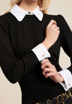 Kobieta w czarnym swetrze z długim rękawem, z białym szpiczastym kołnierzykiem i pasującymi białymi mankietami z złotymi spinkami, poprawiająca jeden mankiet.