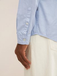 Lichtblauw gestructureerd overhemd met een puntige kraag en knoopmanchetten, gedragen met crèmekleurige broeken. Zichtbare hand die op de broeken rust.