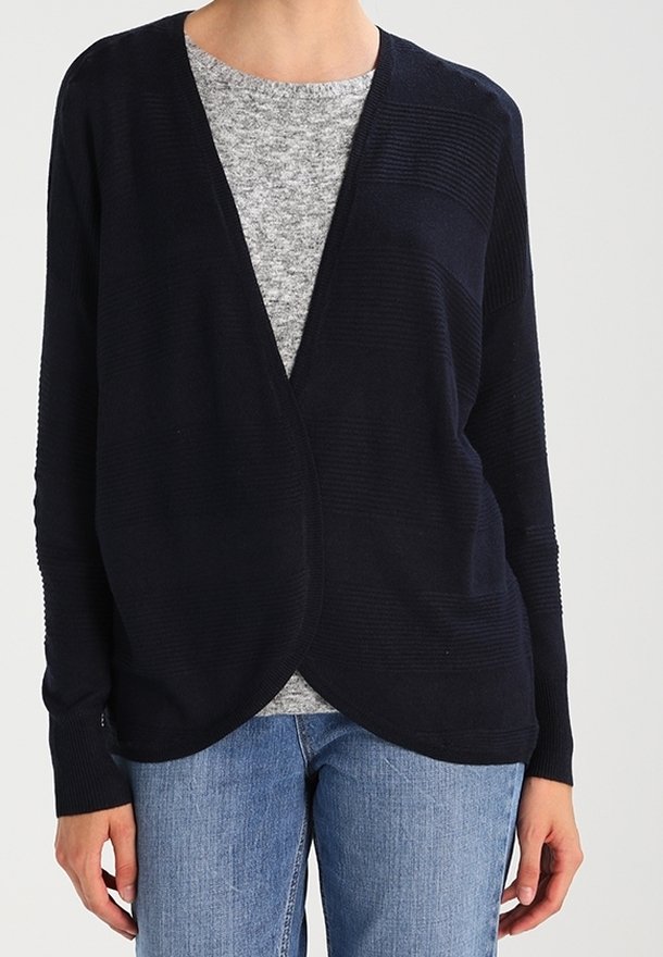 Meriinisininen avointa etuosaa oleva neuletakki, jossa on ribbaustekstuuri, pitkät hihat ja pyöreä helma, jolloin sen päällä on vaaleanharmaa paita, yhdistettynä sinisiin farkkuihin.