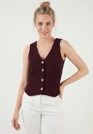 Femeie zâmbitoare cu păr blond prins în coc, purtând o vestă fără mâneci, de culoare mov închis, cu nasturi, și pantaloni albi, stă în picioare pe un fundal simplu.