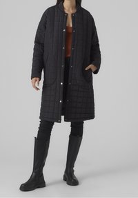 Abrigo largo negro acolchado con cuello redondo, botones a presión y bolsillos laterales, llevado sobre una blusa naranja y combinado con botas de cuero negras.