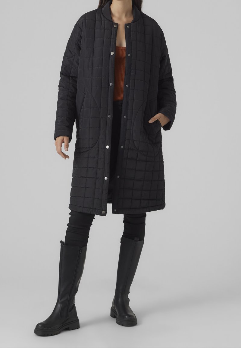 Abrigo largo negro acolchado con cuello redondo, botones a presión y bolsillos laterales, llevado sobre una blusa naranja y combinado con botas de cuero negras.