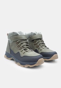 Un par de botas de senderismo verdes y negras con suelas beige, con cordones en la parte delantera y correas ajustables de gancho y bucle sobre un fondo blanco.