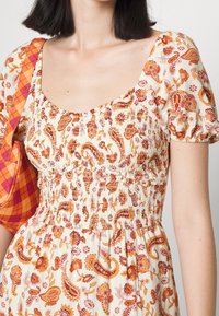 Kleed met bloemendessin in crème met oranje en roze tinten. Heeft een gerimpelde taille en pofmouwen. Vergezeld van een geruit oranje handtas.