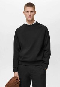 Felpa nera realizzata in morbido materiale, con colletto rotondo, polsini e bordo a coste. Il modello tiene in mano una giacca marrone e indossa pantaloni scuri.