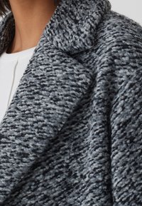 Abrigo de punto gris con una tela suave y texturizada, que presenta un cuello de muesca y un diseño de frente abierto. El patrón incluye diferentes tonos de gris.