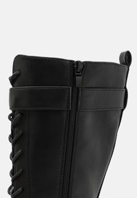 Black leather boot features a textured finish, side zipper, and decorative lacing on the front; sturdy design with a slight sheen.