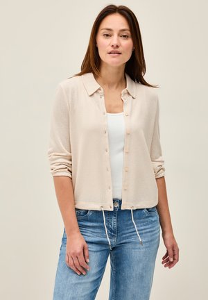 Beige, button-up cardigan gemaakt van een gestructureerd materiaal, met een kraag, omgeslagen mouwen en een trekkoord aan de onderkant. Gemaakt over een wit top.