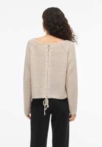 Pull en maille crème avec une coupe courte, doté d'un dos ouvert et d'un détail tressé, associé à un pantalon noir taille haute.