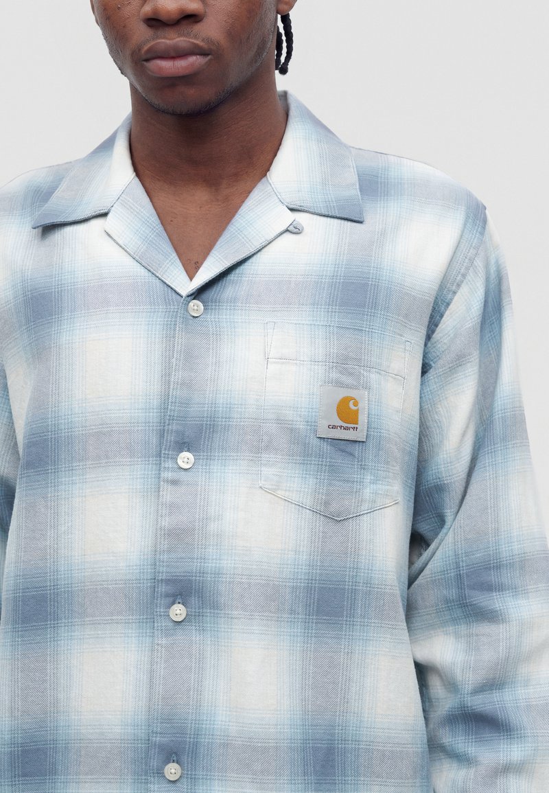 Young man wearing light blue and white plaid button-up shirt with Carhartt logo on left chest pocket, against plain background.